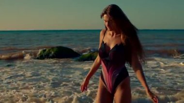 Uzun boylu kız zarif bir şekilde kumsalda gün doğumunda silueti sıcacık bir ışığa vurgu yapıyor. Dalgalar, dingin sabah atmosferini ve gökyüzündeki canlı renkleri izlerken ayaklarını sarıyor.