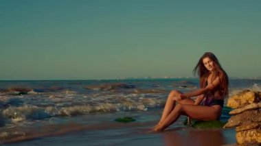 Uzun boylu kız güneş doğarken kayalıklarda zarifçe oturur. Yavaşça ayaklarını dolaştırır. Huzurlu atmosferden ve altın sabah ışığından zevk alır.