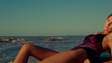 Güzel, uzun boylu bir kız plajda gün doğumunda dinleniyor. Dalgaların sesi ve sabahın erken saatlerindeki hafif parıltı huzurlu atmosferini güçlendiriyor.