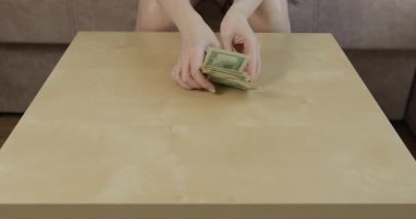 Çağdaş odada kadın eli dikkatle sayıyor. Amerikan dolarlarını pürüzsüz ahşap masa üzerinde yığıyor. Nakitle ilgili detay ve çabalar sergiliyor.