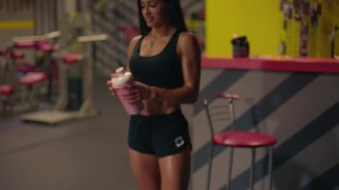 Kız spor salonunda sallanan sudan su içerek antrenmanına hazırlanıyor. Etrafında enerjik parlak renkler ve fitness ekipmanları var..