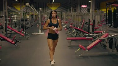 Spor elbiseli bir kız, elinde pembe bir içkiyle spor salonunda yürüyor. Spor salonu, etkin atmosfer yaratan ekipmanlı egzersiz makineleri ve ağırlıklardır..