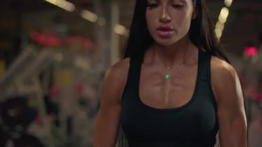 Kız spor salonunda halter kullanarak kaldırma egzersizi yapıyor. Güç eğitimini vurguladığı için dikkati çok yoğun. Spor salonunun arka planında çeşitli spor aletleri var..