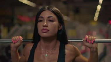 Odaklanmış kız spor salonunda halterle çömelme hareketi yaparak kuvvet antrenmanına başlıyor. Sabah antrenmanlarında azim ve fiziksel kondisyon gösteriyor..