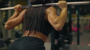 Sporcu kız spor salonunda egzersiz yaparken omuzunda halterle çömeliyor. Vücut şekline ve güç gösterme kararlılığına dikkatle odaklanıyor..