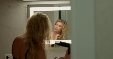 Sarışın kız, aynanın önünde parlak bir şekilde banyoda duruyor. Islak saç kurutma makinesini kuruturken yansımasına bakıyor. Atmosfer rahat ve rahattır.