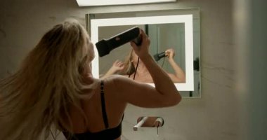 Genç sarışın kız banyoda aynanın karşısında el kurutucusuyla uzun saçlarını kuruturken aynadaki yansımasına odaklanıyor..