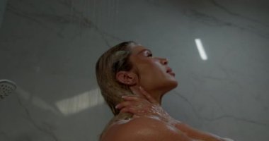 Güzel sarışın kadın duş suyuyla ayakta duruyor, saçlarını ve omuzlarını çağlayarak aşağı iniyor. Uzay zarif mermer duvarlar ve hafif ışıklandırma ile sakin bir atmosfer yaratıyor..