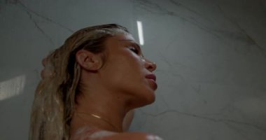 Güzel sarışın kadın duş suyunun içinde etrafından akıyor. Saçları suyla ışıldarken kendine zaman ayırırken rahatlamış görünüyor..
