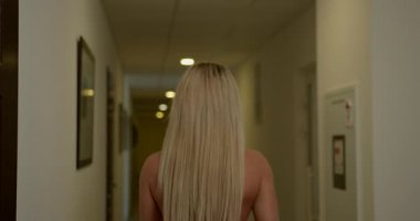 Sarışın kadın güvenli bir şekilde koridorun sonundaki güzel ışıklı koridorda yürür ve kameraya doğru davetkar bir tavırla bakar..