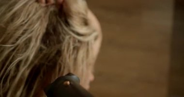 Sarışın bir kız aynanın önünde el kurutma makinesiyle saçını kurutuyor. Banyo atmosferi sabah rutini sırasında rahatlatıcı atmosfer yaratıyor..