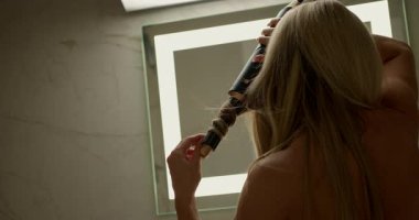 Modern banyodaki kadınlar saç kurutma makinesiyle saçlarını kıvırıyorlar. Yumuşak ışık, saçlarını dikkatlice şekillendirirken rahatlatıcı bir atmosfer yaratır..