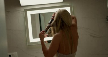 Şık banyodaki kadın saç kurutma makinesinin özel bir eklentisi var. Aynaya bakarken saç stilini mükemmelleştirmeye odaklanıyor..