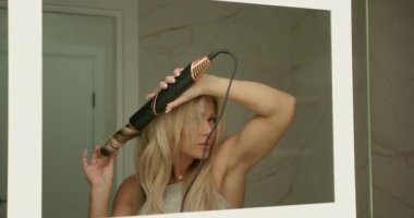 Modern banyoda ayakta duran kadın, saç kurutma makinesinin özel eklenti kıvrımını kullanır ve aynaya bakarken uzun saçlarını perma yapar..