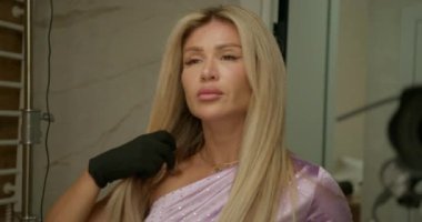 Uzun saçlı bir kadın, saç kurutma makinesiyle buklelerini kıvırıyor ve perma yaptırıyor. Moda tekniğine odaklanmış görünüyor..