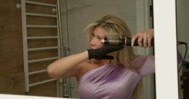 Kadın saç kurutma makinesini kıvırıyor ve perma yapıyor. Zarif mor bir bluz giyerken özel bir eklentisi var. İyi aydınlatılmış banyoda şık bir görünüm elde etmeye odaklanmış..