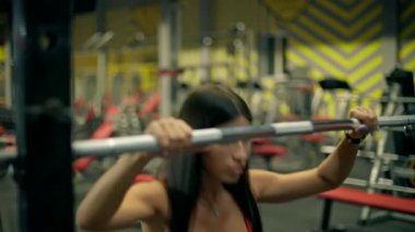 Güzel bir atletik kadın spor salonunda spor salonunda kırmızı bir bluz ve tayt giyerken bir demirci makinesine çömeliyor. Formunu korumaya odaklanıyor..