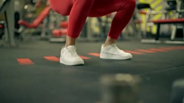 İyi donanımlı spor salonunda kırmızı bluzlu ve tozlu güzel atletik bir kadın demirci makinesine çömeldi. Güç antrenmanlarına ve fitness hedeflerine odaklandı.