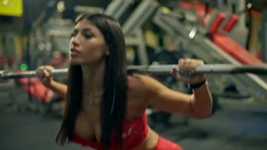 Modern bir spor salonunda odaklanmış kırmızı bluzlu ve tozlu bir kadın düzgün form ve konsantrasyonunu korurken bir demirci makinesine çömelir..