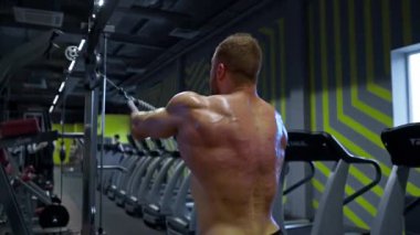 Kaslı bir birey, çağdaş bir spor salonunda kablolu yayında çalışıyor. Çevrede çeşitli egzersiz makineleri ve canlı duvar tasarımları var..