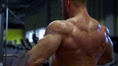 Kaslı adam spor salonunda kablo makinesini kullanarak sırt egzersizine odaklanıyor. Çevre spor deneyimini motive eden spor ekipmanları ve parlak renklerle doludur..