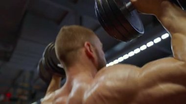 Halterci, bir spor salonunda omuz presi egzersizi yapıyor. Gücünü ve yoğunluğunu gösteriyor. Ayarlar kas tanımını ve ağır ağırlık kaldırma çabalarını vurguluyor..