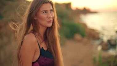 Uzun boylu kız sahilde duruyor güneş doğarken sıcacık altın ışığı manzaranın üzerine saçıyor uzun saçları var rahat bir ifadesi var ve sabahın huzurlu güzelliğinden hoşlanıyor..