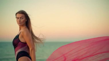 Sersemletici uzun kız plajda gün doğumunda uzun saçları rüzgarda hafifçe savrulurken şafağın canlı renkleri arkasında sakin okyanusu yansıtıyor.