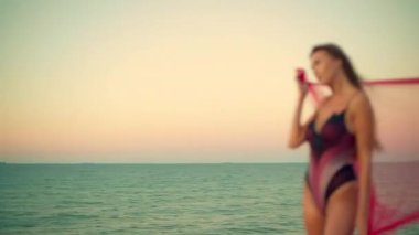 Sakin kumsalda gün doğumunda uzun boylu kız zarif bir şekilde duruş eşarbına sarılmış sakin okyanus sabah güneşinin ılık renklerini yansıtıyor ve sakin bir atmosfer yaratıyor.