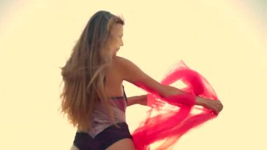 Uzun boylu kız kumsalda gün doğumunda uzun saçları rüzgarda savrulurken gökyüzünün canlı kırmızı eşarp renkleri sahnenin sıcaklığını tamamlıyor.