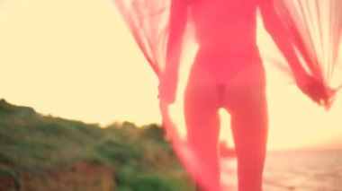 Uzun boylu bir kız kumsalda gün doğumunda elinde akmakta olan peçeyle hafif dalgaları yakalıyor yumuşak dalgalar yavaşça kıyıya vuruyor sakin ve huzurlu bir atmosfer yaratıyor.