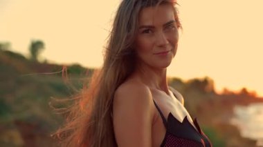 Uzun boylu kız plajda zarif bir şekilde duruyor gün doğumunda uzun saçları hafif bir esintide süzülüyor güneş göğü ılık renklerde boyuyor sakin okyanus dalgaları yavaşça kıyıları sarıyor.