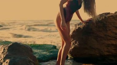 Uzun kız zarif bir şekilde sahilde duruyor gün doğumunda yumuşak dalgalar ayaklarını hafifçe vuruyor altın ışık, dingin okyanus arkaplanına karşı güzelliğini arttırıyor.
