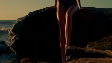 Uzun boylu kız gün doğumunu rahatlatıyor. Kayaların üzerinde oturuyor. Su hafifçe süzülüyor, ayakları şafağın ılık renkleri etrafında huzurlu bir ortam oluşturuyor.