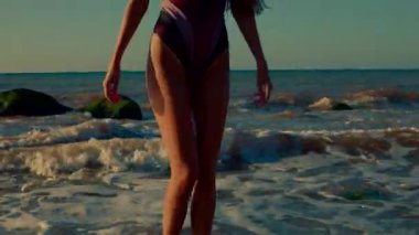 Uzun kız zarif bir şekilde sahilde duruyor. Gün doğumunun huzurunu kucaklıyor. Gökyüzü sıcacık renkleri değiştirirken ayaklarının üzerinde yumuşak dalgalar var.