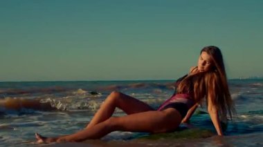Güzel uzun kız gün doğumunda kayalık sahilde dinleniyor. Sakin ifadesi ve yumuşak ışığı dalgaların çarpmasıyla huzurlu bir atmosfer yaratıyor.