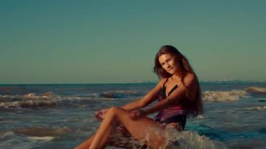 Uzun boylu bir kız kumsalın kenarına oturur güneşin sıcağında güneşlenir nazik dalgalar onu sarar barışçıl bir atmosfer okyanusu yaratır