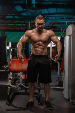 Kaslı adam spor salonunda özgüvenle duruyor ve fiziğinin iyi tanımlandığını vurguluyor. Güç ve kendini adamışlığı vurgulayan, etrafını saran spor ekipmanlarıyla spor yapıyor..