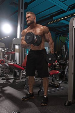 Sporcu adam spor salonunda dambılları kaldırıyor. Çalışmalarına odaklanmış, çeşitli egzersiz ekipmanlarıyla çevrelenmişken kendini adamış ve gücünü sergiliyor..
