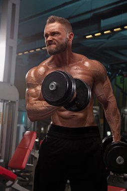 Atletik adam, modern spor salonunda ağır dambılları kaldırırken kaslı vücudunu gösteren güçlü bir antrenman seansına katılır. Atmosfer enerjik ve yoğun..