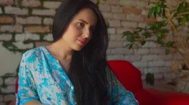 Kameraya uzun siyah saçlı kız görünüşünü tamamlayan renkli bir bluz giyiyor. Arka planda bir tuğla duvar ve sıcak atmosferi geliştiren yeşillik yer alıyor..