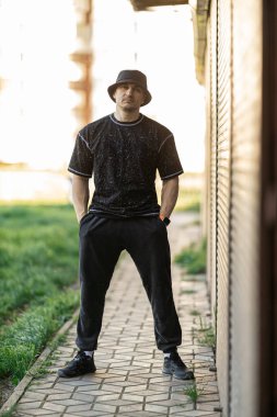 Genç adam, açık havada bir duvarın yanında rahat bir şekilde siyah bir kıyafetin içinde duruyor. Öğleden sonranın ılık ışığı, onun tarzını ve davranışlarını vurgulayan sahneyi güçlendiriyor..