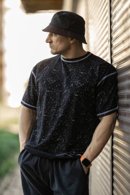 Adam, yumuşak bir akşam ışığında dokulu bir duvara karşı duruyor. Siyah bir kısa kollu gömlek ve bol pantolon giyiyor. Kova şapka ve saat takıyor..