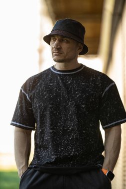 Adam kentin bir köşesinde siyah bir kova şapka ve desenli siyah bir gömlek giyerek duruyor..