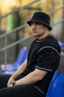 Adam mavi tribünde tek başına oturmuş siyah desenli gömlek ve kova şapka giyiyor. Etrafı sporlarla çevrili olan mesafeye dikkatle bakıyor..