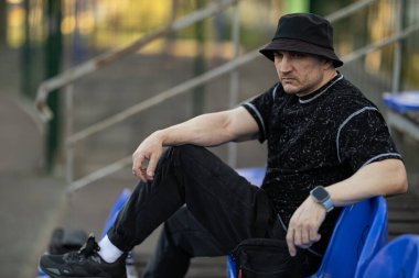 Adam çocuk parkında rahat bir şekilde oturur. Koyu renk kıyafetler giyer. Mavi koltukta dinlenir ve düşünceli bir şekilde öğleden sonra güneşine bakar..