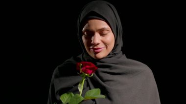 Siyah tesettürlü kadın sessizce duruyor. Gözleri kapalı. Nazikçe kırmızı gülü tutuyor ve kokluyor. Geçmişi karanlık, onun barışçıl ifadesini ve bağlantı çiçeğini vurguluyor..
