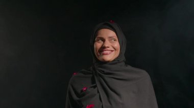 Belirsiz ışıklandırılmış bir çevrede tesettürlü kırmızı aksanlı kadın sıcak bir şekilde gülümser. Neşeli ifadesi, çeşitliliği ve toplumu kutlayan kültürel etkinliğin canlı atmosferini yansıtıyor..