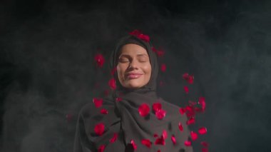 Siyah tesettürlü kadın, gözleri dingin bir şekilde kapalı dumanlı arka planda dalgalanan kırmızı gül yaprakları gibi, huzurlu bir atmosfer yaratıyor..
