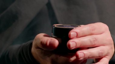 Tesettürlü kadın, öğleden sonra evde otururken elinde küçük bir konteynır tutarken görülüyor. Ilık ışık etrafında rahat bir atmosfer yaratıyor..